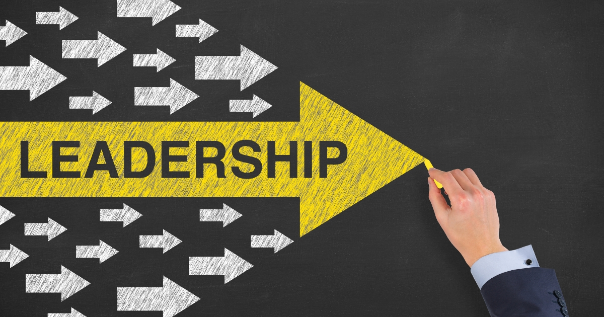 Yellow arrow labeled LEADERSHIP being pulled forward by a hand, cutting through a field of white arrows on a chalkboard background.